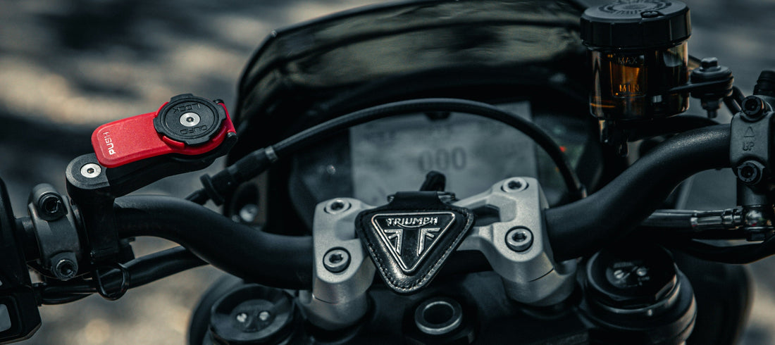 Close-up of a motorcycle handlebar area with a visible 5" carplay screen, black tank, and red handbrake lever, photographed outdoors on a blurred path.
