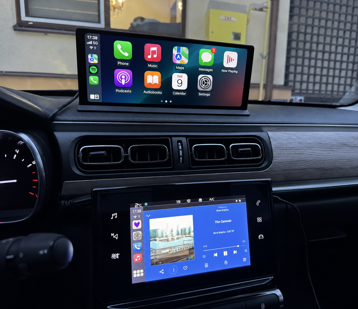 A car dashboard with two touchscreen infotainment displays showing Apple CarPlay and Android Auto interfaces, photographed in a real vehicle interior.